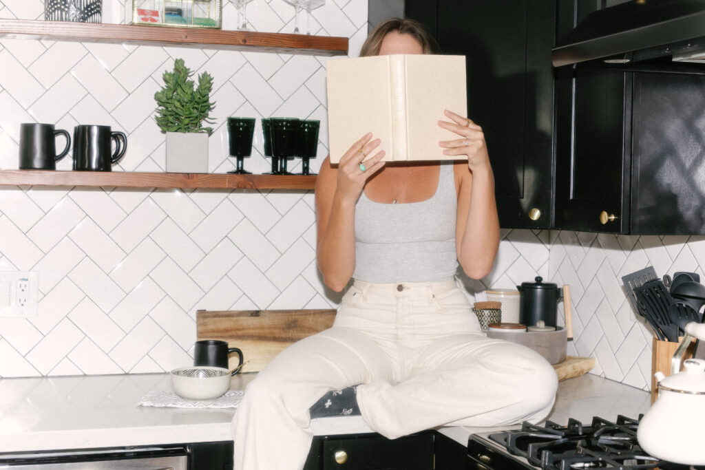 A small business copywriter sits on a kitchen counter, hiding her face with a book.