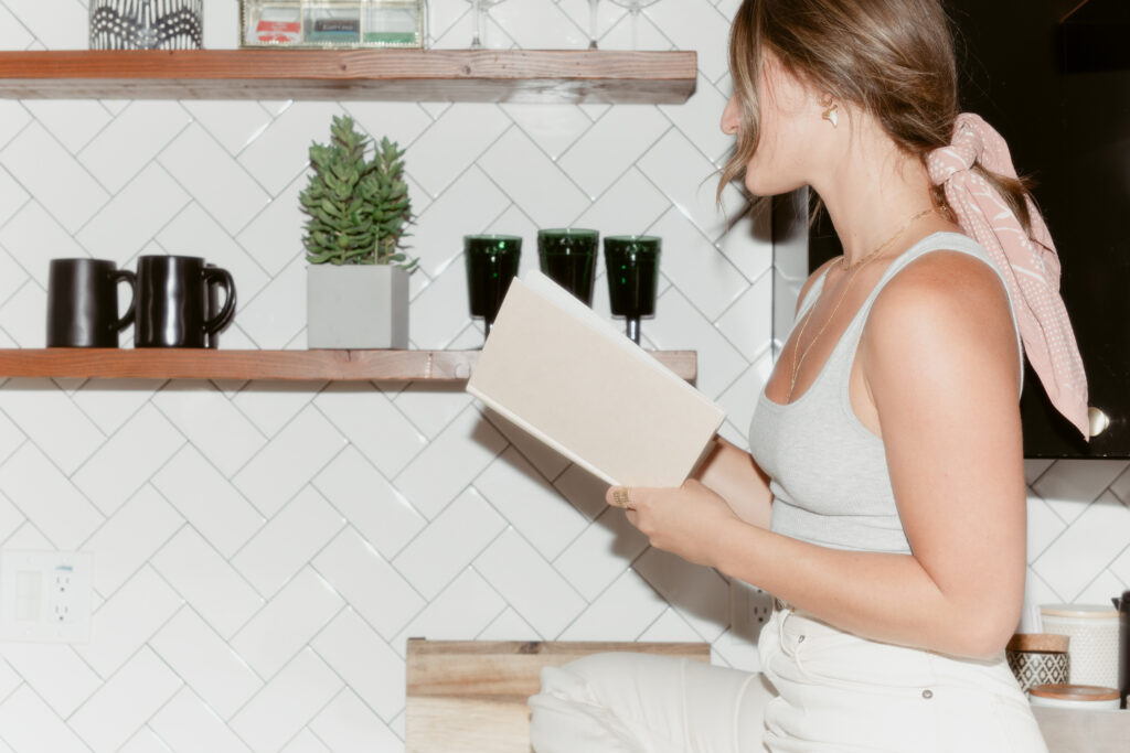 A copywriter for small businesses sits on a kitchen counter in an all-white room, reading a copywriting book.