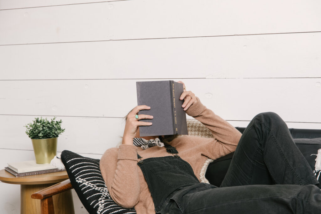 A small business blog writer reads a book on a couch, hiding her face with the book.