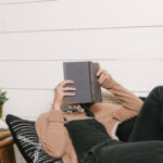 A small business blog writer reads a book on a couch, hiding her face with the book.